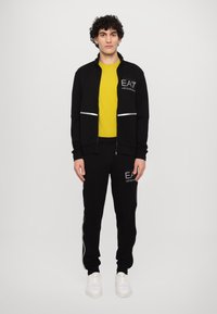 Black zip-up jacket with silver stripes and logo, paired with a yellow t-shirt and black pants. White sneakers with textured soles.