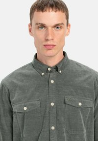 Grüne Cordhemd mit Button-Down-Kragen, ausgestattet mit zwei Vordertaschen und hellen Knöpfen. Strukturierter Stoff mit feinem Muster.
