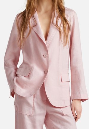 Vrouw die een lichtroze linnen blazer draagt met twee knopen en bijpassende broek met grote voorzakken, staand met één hand in de zak.
