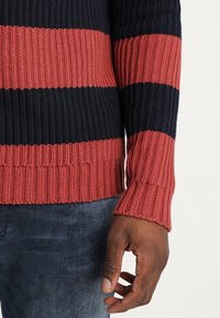Knitted sweater with horizontal stripes in navy blue, brick red, and maroon. Features ribbed cuffs and hem, showcasing a textured surface.