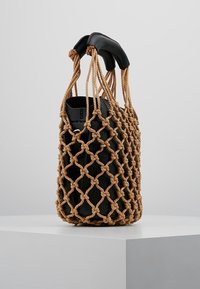 Tkaná taška s uzlovým lanovým vnějším povrchem v hnědé barvě, s hladkým černým interiérem a černými koženými uchy. Cylindrický tvar.