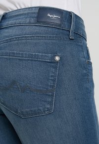 Jeans in denim blu con un'etichetta in pelle marchiata, dettagli sulla tasca posteriore con cuciture arcuate e accessori in metallo argento.