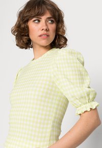 Top com mangas bufantes amarelo claro, com um padrão texturizado e quadriculado. Apresenta um decote redondo e punhos com babados. Design justo.