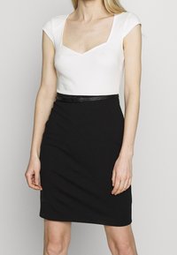 Robe ajustée avec un corsage blanc à encolure dégagée et une jupe noire arrivant au genou. Comprend une ceinture noire fine à la taille. Texture lisse.