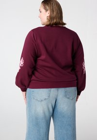 Bordeauxkleurige pullover met ballonmouwen en roze bloemenborduursel op de linker mouw, gecombineerd met lichtblauwe wijd uitlopende jeans.