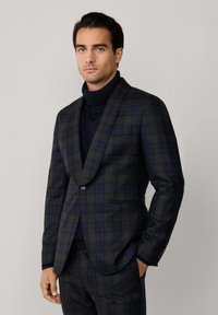 Uomo che indossa un abito a quadri verde scuro e navy con giacca a singolo bottone e maglione a collo alto navy, in piedi contro uno sfondo chiaro e uniforme.