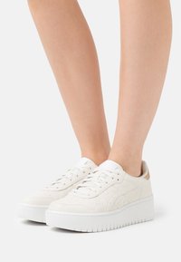 Persona che indossa sneakers platform bianche a basso profilo con chiusura a lacci su uno sfondo bianco, mostrando i piedi e le gambe inferiori.
