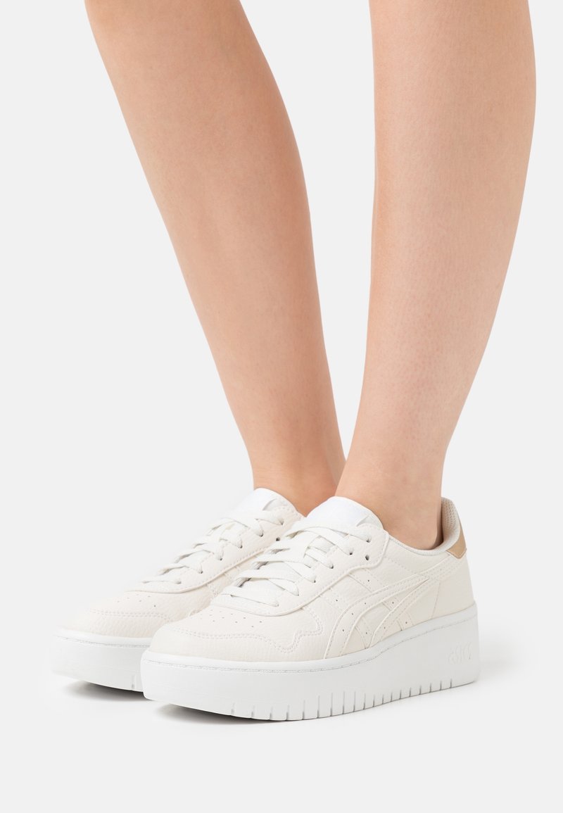 Persona che indossa sneakers platform bianche a basso profilo con chiusura a lacci su uno sfondo bianco, mostrando i piedi e le gambe inferiori.