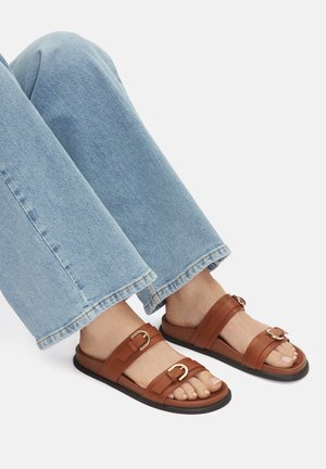 Brązowe skórzane sandały z dwiema szerokimi paskami i metalowymi klamrami, w połączeniu ze jasnoniebieskimi dżinsami w stylu flare z surowym wykończeniem dołu.