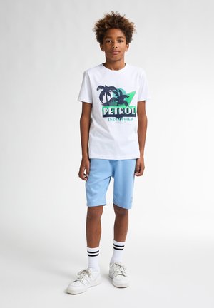 Weißes T-Shirt mit einem grünen und blauen Grafikdesign, das Palmen zeigt, kombiniert mit hellblauen Shorts und weißen Sneakers mit schwarzen Streifen auf den Socken.