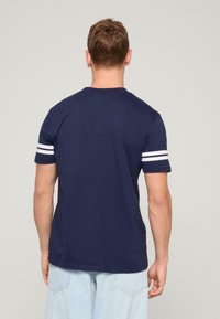 Camiseta de algodón de color azul marino con rayas blancas en las mangas cortas. Diseño clásico de cuello redondo. Textura suave, corte regular.