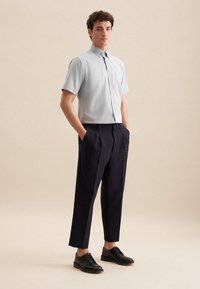 Camicetta azzurra a maniche corte con bottoni e tasca, abbinata a pantaloni scuri a pieghe color navy e scarpe eleganti nere, in piedi contro uno sfondo neutro.