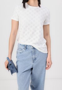 Vit kortärmad T-shirt med utskärningsmönster, kombinerad med ljusblå högmidjade denimjeans. Modell som håller en liten blå handväska.