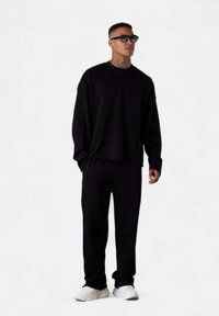 Sweatshirt noir à manches longues et col rond associé à un jogging assorti, fabriqué dans un tissu doux. Des baskets blanches complètent la tenue.