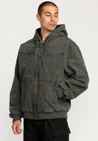 Grey canvas jacket with a hood, featuring a front zipper, drawstring, two front pockets, and ribbed cuffs and hem. Accented with a logo patch.