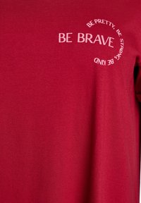 Red fabric with white text reading "BE BRAVE" and circular text "BE PRETTY, BE STRONG, BE KIND" on chest area.