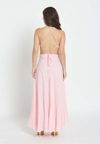 Mujer con cabello largo y oscuro, vestida con un vestido maxi rosa claro de espalda descubierta con tirantes cruzados y tacones altos amarillos, de pie contra un fondo blanco.