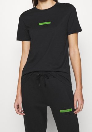 Vrouw draagt een zwart Calvin Klein T-shirt en zwarte sweatpants, beide met kleine groene rechthoekige logopatches.