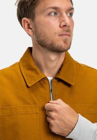 Giacca color senape con colletto, zip frontale e due grandi tasche. Presenta un tessuto testurizzato e dettagli di cucitura visibili.