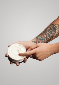Crema bianca in un barattolo rotondo tenuto in due mani, con un dito che affonda nella crema; braccio tatuato visibile. Texture liscia, superficie del barattolo opaca.