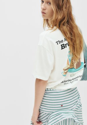 Giovane donna con lunghi capelli rossi indossa una t-shirt grafica bianca con un disegno di pane e latte, abbinata a una gonna a righe verdi, su sfondo bianco.