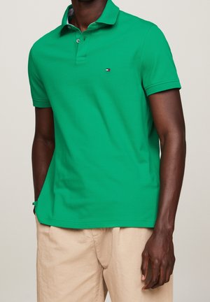 Man draagt een felgroen poloshirt met korte mouwen en een klein rood-wit-blauw logo op de borst, gecombineerd met beige broek.