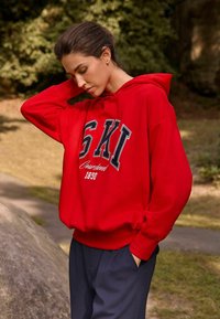 Sudadera con capucha roja con un gran gráfico negro "SKI" y texto blanco. Tela suave, corte holgado, puños acanalados, combinada con pantalones azul marino oscuros.