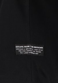 Etiqueta de tela negra con texto blanco que dice "ESCAPE DE / LO DESCONOCIDO" y detalles adicionales de la marca. El texto está impreso sobre una superficie texturizada.