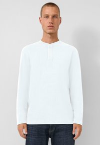 Junger Mann mit kurzen Haaren, der ein weißes Langarm-Henley-Shirt und eine dunkelblaue Jeans trägt und vor einem schlichten hellgrauen Hintergrund steht.