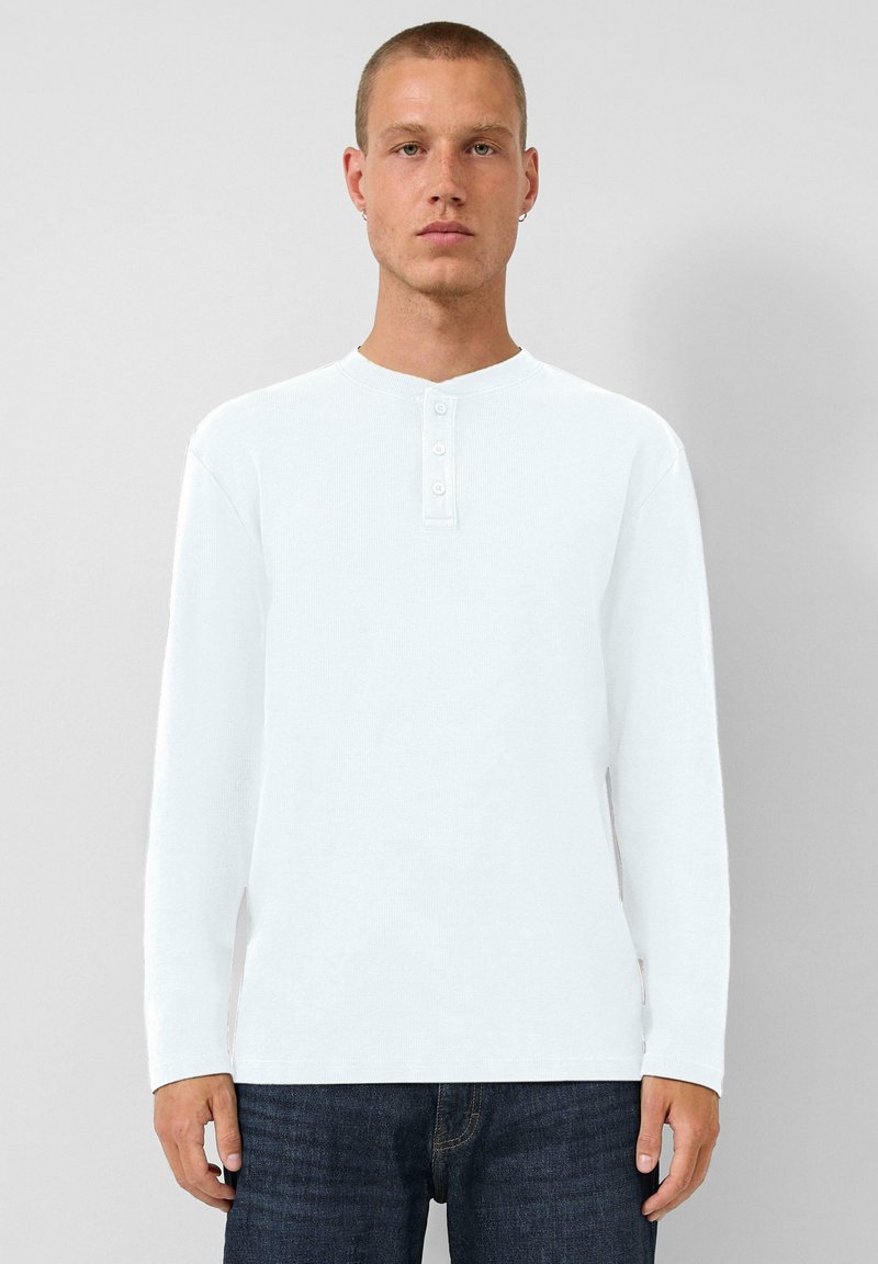 Junger Mann mit kurzen Haaren, der ein weißes Langarm-Henley-Shirt und eine dunkelblaue Jeans trägt und vor einem schlichten hellgrauen Hintergrund steht.