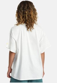 Camiseta blanca de gran tamaño con mangas cortas y un corte holgado; textura suave. Acento de texto amarillo en la manga. Diseño minimalista, sin patrones.