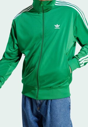 Persona que lleva una chaqueta deportiva verde de Adidas con rayas blancas en las mangas y vaqueros azules, con las manos en los bolsillos de la chaqueta.