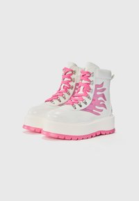 Witte platformlaarzen met een roze vlamontwerp, textuurleer, roze veters en zilveren hardware. Dikke witte zool met roze accentuitgangen.