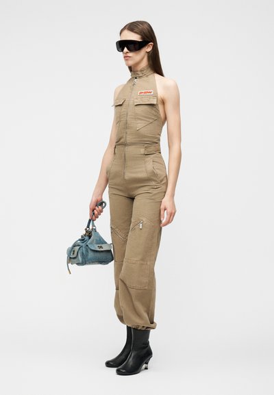 Femme portant une combinaison beige sans manches avec une fermeture éclair à l'avant, des bottines noires, de grandes lunettes de soleil noires, tenant un petit sac à main bleu.