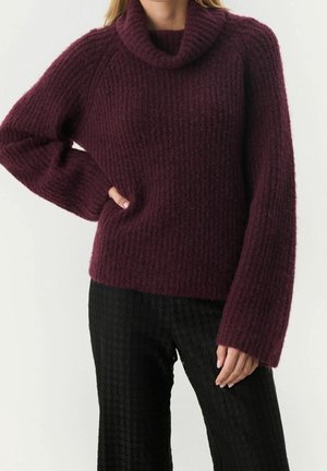 Femme portant un pull à col roulé côtelé violet foncé avec des manches longues et amples et un pantalon noir texturé, posant avec une main sur la hanche.