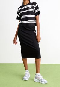 Schwarz-weiß gestreiftes, gekürztes T-Shirt mit PUMA-Logo, kombiniert mit einem schwarzen, gerippten Midirock und hellen Sportsneakern. Grüner Hintergrund.