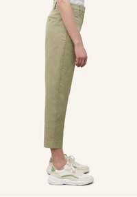 Personne portant un pantalon court taille haute vert clair et des baskets blanches avec des accents verts, debout de profil sur un fond uni.