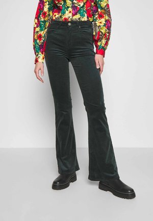 Žena nosící tmavě zelené manšestrové rozšířené kalhoty, černé masivní boty a barevnou květinovou blůzu s dlouhým rukávem, stojící před jednoduchým pozadím.