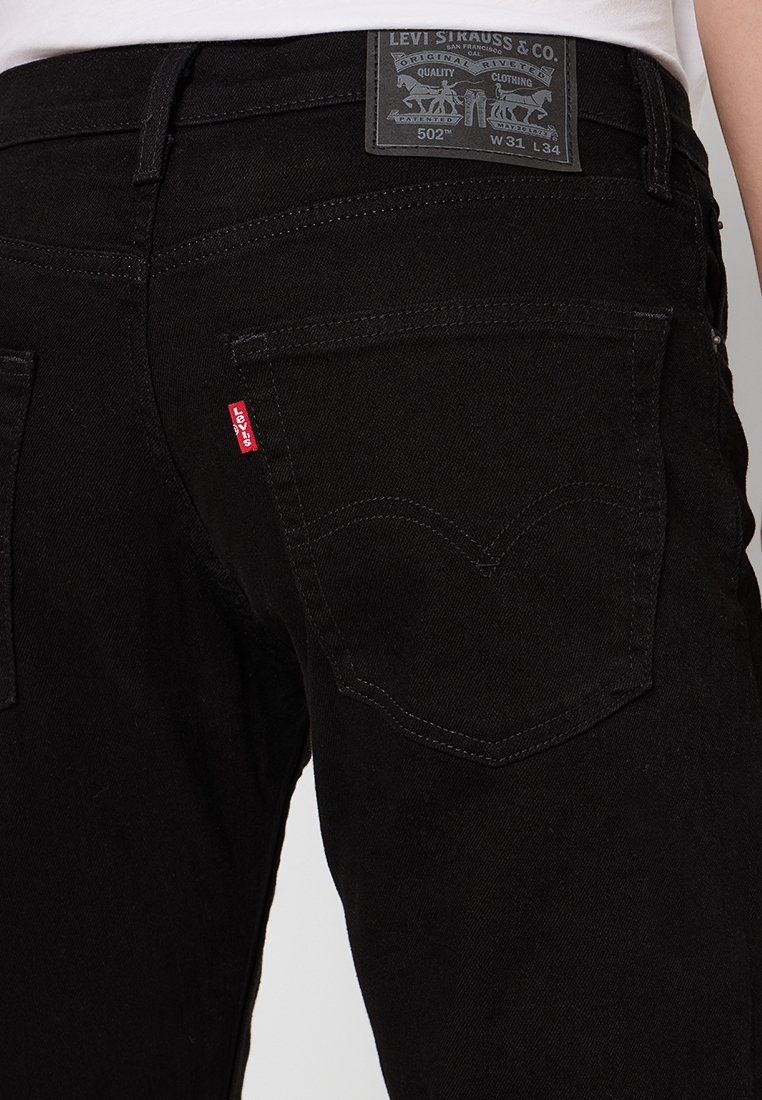 Schwarze Denim-Jeans mit einem Etikett am Bund. Verfügt über runde Gesäßtaschen und einen roten Tabakzent. Genähte Details an den Taschen.