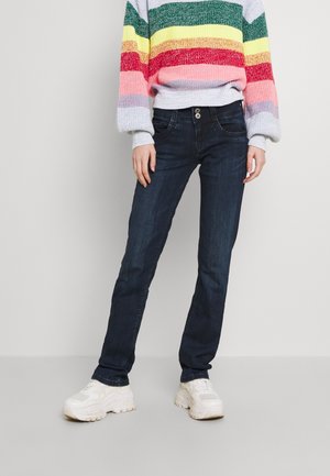 Persoon die donkerblauwe spijkerbroek, witte chunky sneakers en een kleurrijke gestreepte trui met groene, gele, rode, roze en grijze strepen draagt.