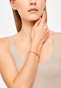 Kvinde iført en beige tanktop, der rører ved sin nakke, og viser et armbånd i rosaguldsfarve med hjerteformede led på håndleddet.