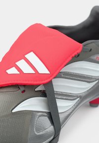 Botín de fútbol gris con una superficie texturizada, una correa roja brillante con detalles en blanco y un diseño aerodinámico elegante.