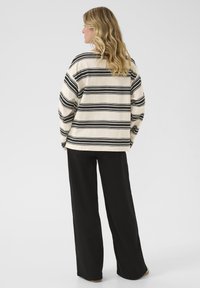 Gestreifter, cremefarbener Pullover mit schwarzen horizontalen Linien, lockerer Schnitt, weite Ärmel. Kombiniert mit schwarzen weiten Hosen. Rückansicht. Glatter Stoff. 