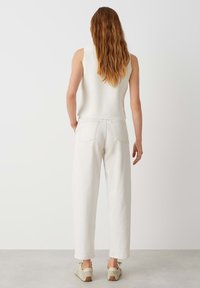 Un haut sans manches blanc associé à un pantalon large écru, avec des poches à rabat et des chaussures de teinte neutre, vu de l'arrière.