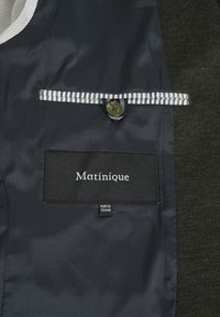 Black fabric tag with white text "Matinique" on a dark green inner lining, striped accents, and a button closure. Contains size labels.