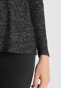 Svart och grå melerad långärmad topp med rundad nederkant och mjuk textur, kombinerad med mörka jeans. Ärmen slutar vid handleden.