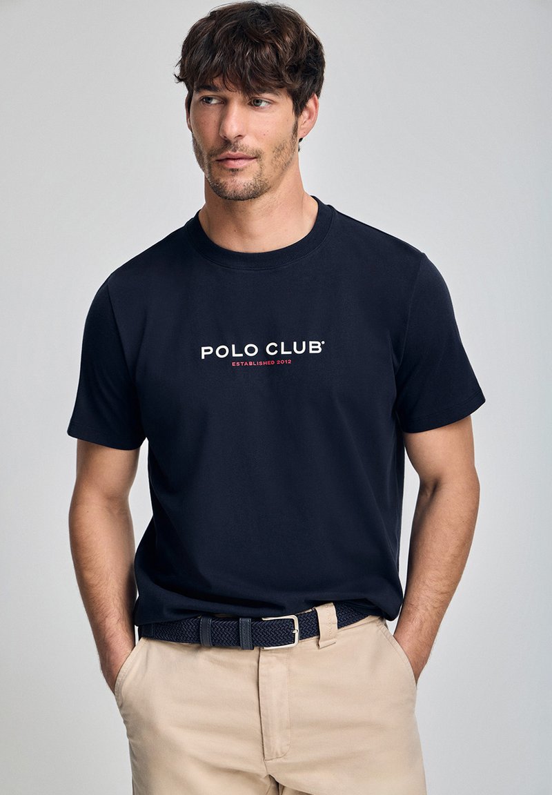 Navyblå bomullströja med texten "POLO CLUB ETABLERAD 2012" tryckt i vitt och rött på bröstet. Rund halsringning och korta ärmar.
