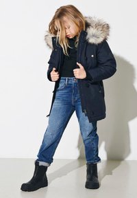 Marinblå vinterparka med huva av fuskpäls, dragkedja och knäppning med knappar. Matchad med ljusblå jeans och svarta vattentäta ankelboots.