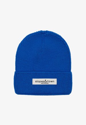 Blauwe gebreide beanie met een geribde omslag. Voorzien van een wit label met "slopes&town AMSTERDAM." Soepele textuur, rond ontwerp aan de bovenkant.