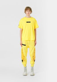 Camiseta de algodón amarilla con un parche de logo negro, combinada con joggers amarillos a juego que presentan acentos de diseño intrincados y detalles negros.